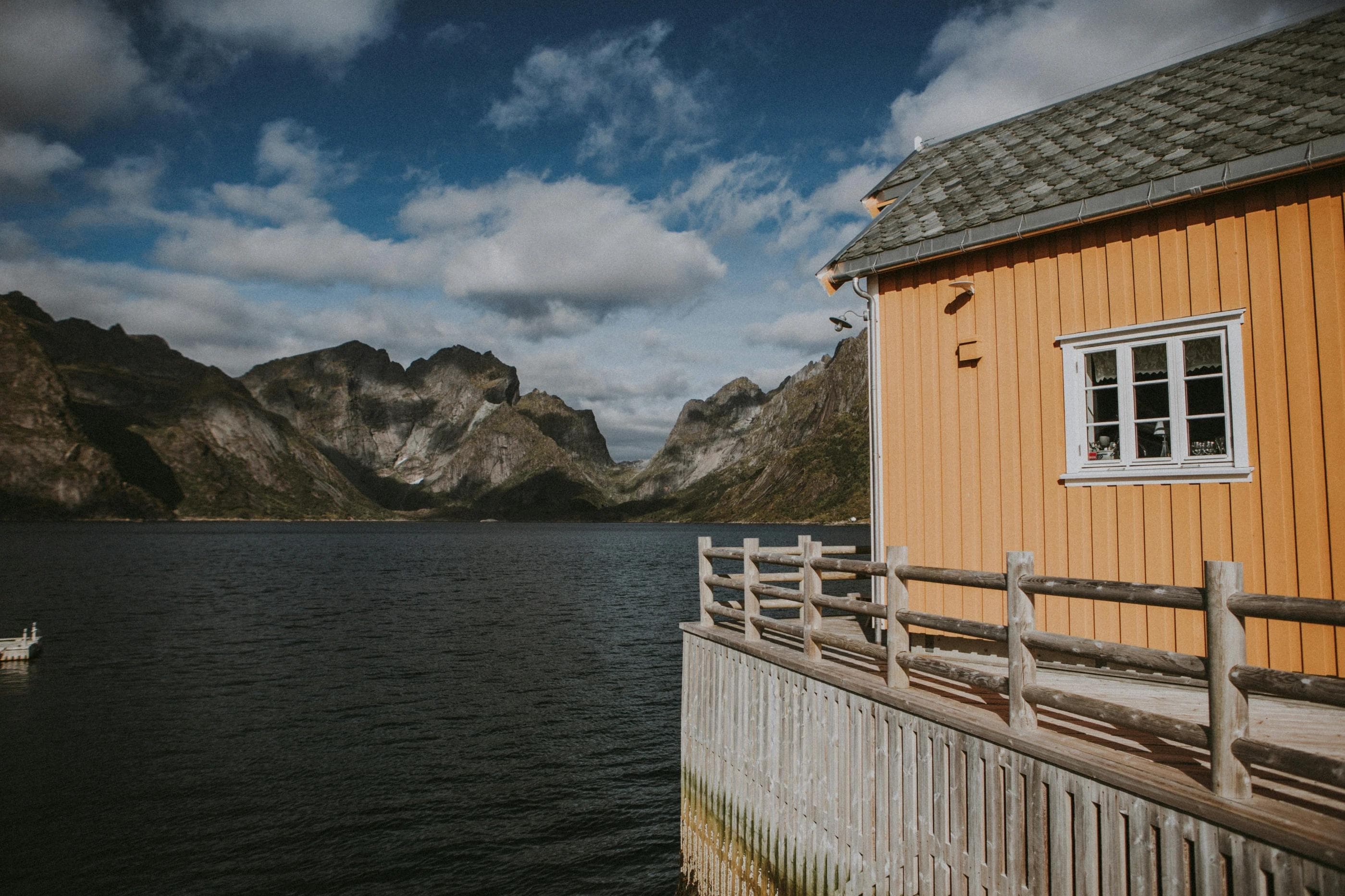 Et gult naustaktig trehus står på påler i sjøkanten, med utsikt mot dramatiske, spisse fjellformasjoner.